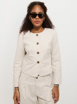 A person with long curly hair wears dark sunglasses and the Soho Jacket In Cotton Linen with matching pants, standing against a plain white background.