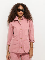 A person with curly hair wears black sunglasses and a light pink Painter Jacket In Herringbone with large front pockets and rolled-up sleeves, paired with matching pink pants, against a plain white background.