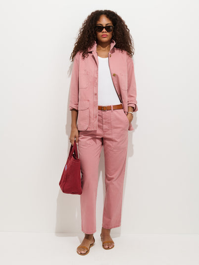 A woman with curly hair, sunglasses, a pink Neil Pant In Herringbone, matching jacket, white top, sandals, and a red bag stands against a white background.