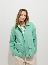 A woman with light brown hair wears the Painter Jacket In Herringbone, an oversized mint green cotton piece, over a black-and-white striped shirt and light beige pants, standing against a plain white background.