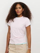 A woman with curly brown hair stands against a white background wearing the Prospect Tee In Cotton Jersey, a light pink women’s designer T-shirt layered over a light blue top and beige pants. She has a neutral expression and simple accessories.