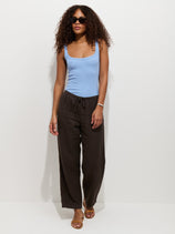 A woman with curly hair, black sunglasses, a light blue sleeveless top, sandals, bracelet, and wide-leg Hudson Pant In Linen with a pull-on waist stands against a plain white background.