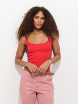 A person with long curly hair wears the 365 Ribbed Tank in red with pink high-waisted pants, standing against a white background and looking at the camera with a neutral expression.