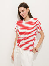 A woman with straight brown hair wears the Garcon Striped Tee In Linen Cotton, featuring red and white stripes, paired with tie-waist white pants. She stands against a plain white background with a neutral expression.