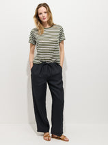 A person with medium-length blond hair stands against a white background wearing a green and white striped T-shirt, brown sandals, and the Hudson Pant In Linen with a pull-on waist. Their hands are in their pockets.