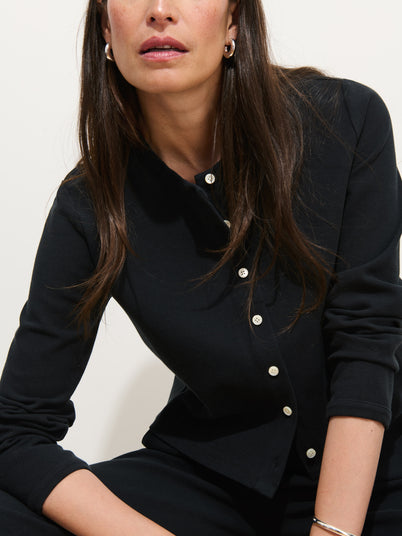 A woman with long brown hair wears the Coco Sport Cardigan, featuring troca shell buttons, paired with silver hoop earrings. She is seated with one arm on her leg, facing slightly to the side against a plain light background.