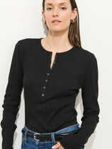 A woman in the Laine Henley In Fine Rib, a black cotton-modal long-sleeve top, pairs it with blue jeans and stands against a white background with a neutral expression.