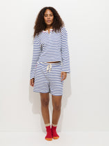 A woman with curly hair wears The Striped Waffle Short and a matching blue and white striped long-sleeve shirt, inspired by Alex Mill pajamas, paired with red socks, against a plain white background.