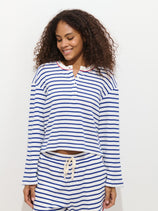 A person with curly hair smiles and looks down while wearing The Striped Waffle Sleeping Henley—a blue-and-white striped long-sleeve top with drawstring shorts. The background is plain white.