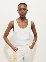 A person stands against a plain white background, wearing the Garcon Tank In Linen Cotton with white pants—one hand in their pocket, the other holding a beige netted bag.