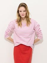 A woman with long, straight light brown hair wears the Avignon Striped Tee in Cotton—white with pink Breton stripes—paired with a knee-length red skirt, standing against a plain white background and looking at the camera.