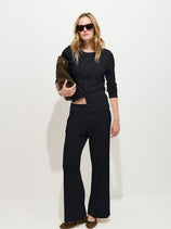 A model wears a black button-up long-sleeved top and garment-dyed black Scout Pull On Pant in Cotton Rib with an elastic waistband, while holding a dark brown clutch.