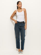 A woman stands against a white background, wearing The Carla High Rise Straight—dark blue, non-stretch cotton denim jeans with a black belt, a white tank top, and silver ballet flats. She gazes to the side with one hand near her pocket.