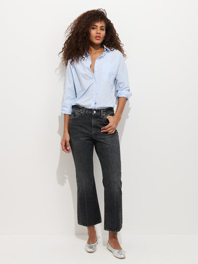 A woman with curly hair wears a light blue button-up shirt, Bessette Mid Rise Boot Cut Jean, and silver flats, standing against a plain white background.
