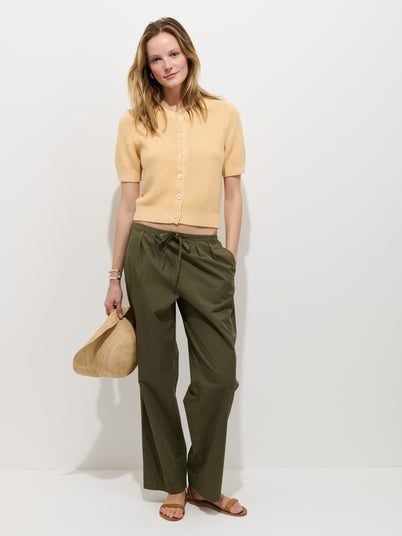 A woman stands against a white wall in the Hudson Pant In Cotton Twill—olive green, wide-leg style—paired with a beige short-sleeve cardigan, brown sandals, and holding a straw hat, exuding relaxed and casual vibes with a slight smile.