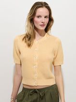 A woman with light brown hair models the Elle Cardigan In Linen Cotton, a short-sleeve, hand-finished piece with Troca shell buttons, paired with olive green drawstring pants against a plain white background.