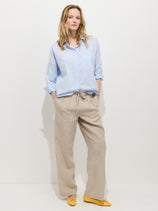 A woman stands against a white background wearing a light blue button-up shirt and the Hudson Pinstripe Pant in Linen, featuring a wide-leg cut and pull-on waist. She completes her look with yellow shoes, hands in her pockets and a relaxed expression.
