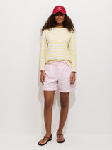 A person in a red baseball cap, yellow and white striped long-sleeve shirt, Lucie Short In Linen Cotton (light pink, pleated with elastic waistband), and black sandals stands against a plain white background.