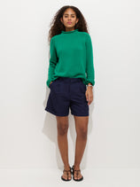 A person with curly hair stands against a white background wearing a green long-sleeve sweater, the Lucie Short In Linen Cotton in navy blue with an elastic waistband and pleats, and black sandals. Their hands are in their pockets.