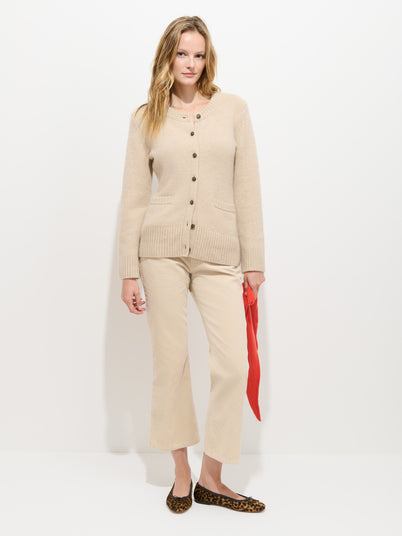 A woman stands against a white background wearing the Bessette Mid-rise Jean in Corduroy and a beige cardigan, holding a red scarf in her left hand and finishing the look with leopard-print flats.