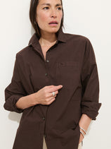 A person with long dark hair, silver jewelry, and a neutral expression stands against a plain light background, wearing the Jo Shirt in Cotton Poplin—a brown 100% cotton button-up with Troca shell buttons—and touches the shirt pocket.