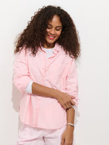 Smiling and looking down, a person with curly hair wears the Jo Shirt in Cotton Poplin—a light pink 100% cotton button-down—layered over a white top with matching light pink pants, set against a plain white background.