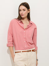 A woman with straight brown hair wears the Jo Shirt In Cotton Poplin, a loose-fitting pink button-down with rolled-up sleeves, paired with beige high-waisted pants and a brown belt, standing against a plain white background.
