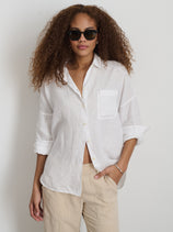 A woman with curly hair wears dark sunglasses, light beige pants, and The Jo Shirt In Linen—a loose 100% linen shirt with real shell buttons and rolled sleeves—while standing against a plain white background.