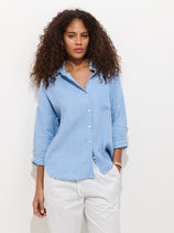 A woman with long curly hair stands against a white background, wearing The Jo Shirt In Linen in light blue paired with white pants. The relaxed-fit shirt has rolled-up sleeves, and she looks comfortable and at ease.