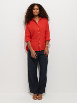 A woman with curly hair stands against a white background, wearing a loose red shirt, tan sandals, and the Hudson Pinstripe Pant In Linen—navy blue wide-leg pants with a pull-on waist. She looks at the camera with a relaxed expression.