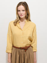 A woman with light brown hair wears The Jo Shirt In Linen, a relaxed fit 100% linen shirt with real shell buttons, tucked into a brown skirt with a braided belt, posed against a plain white background.