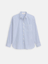 The Jo Striped Shirt In Cotton Poplin, featuring blue and white vertical stripes, long sleeves, front pocket, collar, and French seams, is crafted from 100% cotton and shown laid flat on a plain white background.