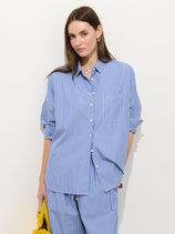 A woman with long brown hair wears the Jo Striped Shirt in Cotton Poplin—loose-fitting, blue and white stripes—with matching pants, standing against a white background and holding a yellow bag.