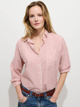 A person with long, light brown hair wears the Jo Striped Shirt In Cotton Poplin—red and white stripes—paired with blue jeans and a brown belt, standing confidently against a plain white background.
