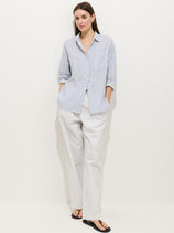 A woman with a neutral expression stands against a white background wearing the Hudson Pant In Cotton Twill, paired with a light blue and white striped shirt and black flat sandals, her hands in her pockets.
