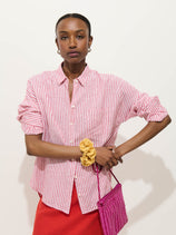 A person wears the Jo Striped Shirt In Linen (red and white stripes) with a red skirt, standing against a plain background, holding a pink woven handbag and a yellow scrunchie on their wrist.