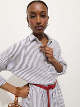 Someone stands against a white background wearing the Jo Striped Shirt In Linen, a relaxed fit, long-sleeved, white and gray striped 100% linen dress with real shell buttons and a red braided belt, holding a brown beaded bag.