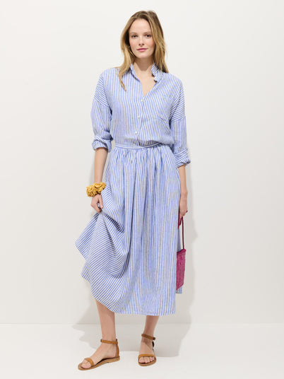 A woman stands against a plain background wearing the Sarah Striped Skirt In Linen with a matching blue and white striped linen button-down shirt, tan sandals, a pink purse, and a yellow scrunchie on her wrist.