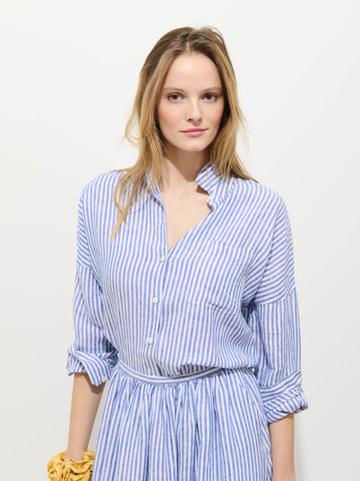 A woman with long blond hair wears the Jo Striped Shirt In Linen—a relaxed fit blue and white striped dress with rolled-up sleeves and a cinched waist—against a plain white background, holding a yellow scrunchie.