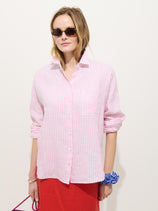 A woman in sunglasses wears the Jo Striped Shirt In Linen—pink and white stripes, real shell buttons, collar popped—paired with a red skirt and blue scrunchie, standing against a plain white background.