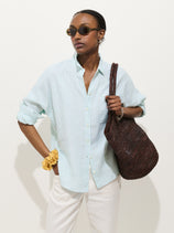 Wearing sunglasses and the Jo Striped Shirt In Linen with real shell buttons, white pants, a large brown woven bag, and a yellow scrunchie on their wrist, this person stands against a plain background.