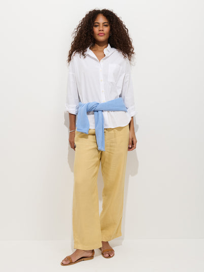 A person stands against a plain white background wearing a white button-up shirt, the Hudson Pant In Linen (wide leg with pull-on waist), sandals, and a light blue sweater tied around their waist.
