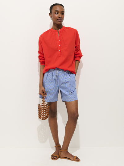 A person stands against a white background wearing a bright red long-sleeve shirt, Hudson Striped Short In Poplin, brown sandals, and holding a small woven bag.