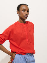 Wearing the Nina Popover in bright red cotton voile with white buttons, paired with blue-and-white striped pants, a person stands against a plain white background holding a brown beaded strap.