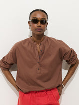 Against a white background, a person wears dark sunglasses, bright red pants, and the Nina Popover in Cotton Voile—an oversized women's designer shirt with rolled-up sleeves and buttons—standing with hands on hips.