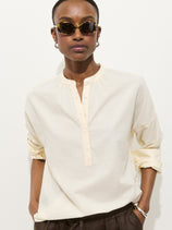A person in sunglasses, dark pants, and the Nina Popover—an oversized women’s designer shirt in cream 100% cotton voile with rolled sleeves—stands against a plain white background.