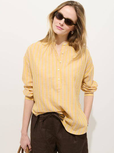 A person stands against a plain light background wearing sunglasses, brown pants, holding a woven bag, and the Nina Striped Popover In Double Gauze—a women's oversized fit shirt in yellow and white striped 100% cotton gauze.