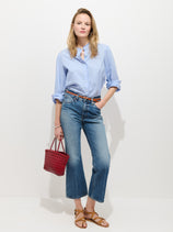 A woman with blonde hair poses against a white background wearing a light blue cotton button-up shirt, Bessette Mid Rise Boot Cut Jeans, brown sandals, and holding a small red woven handbag.