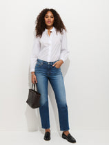 Woman with curly hair stands against a white background wearing a white button-down shirt, Jane Mid Rise Comfort Jean, black loafers, and holding a black woven handbag.