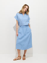 A woman stands against a white background, wearing the garment-dyed Maddie Blouse in light blue (100% linen), tucked into the matching Maddie Skirt In Linen—both in blue—and paired with tan sandals.